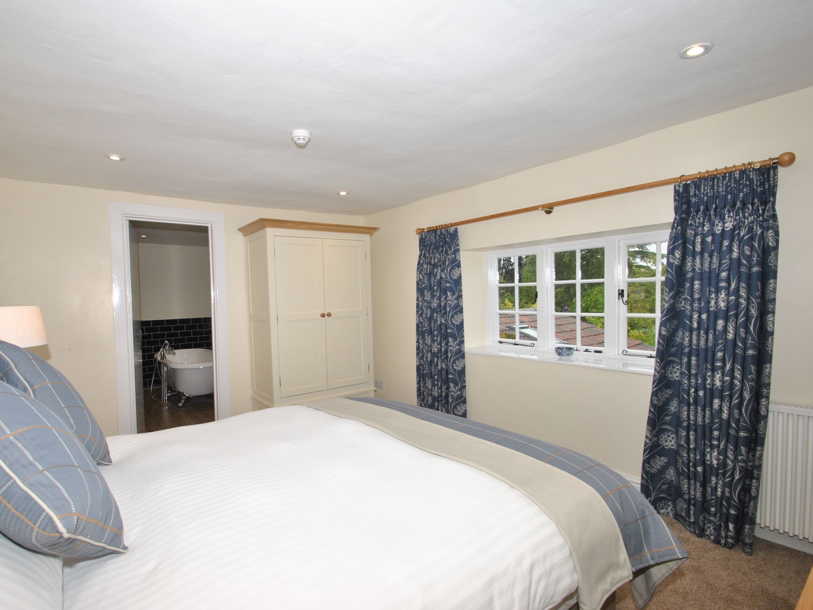 A bedroom with a bed, a wardrobe, a window with curtains, and a bathroom visible through an open door.