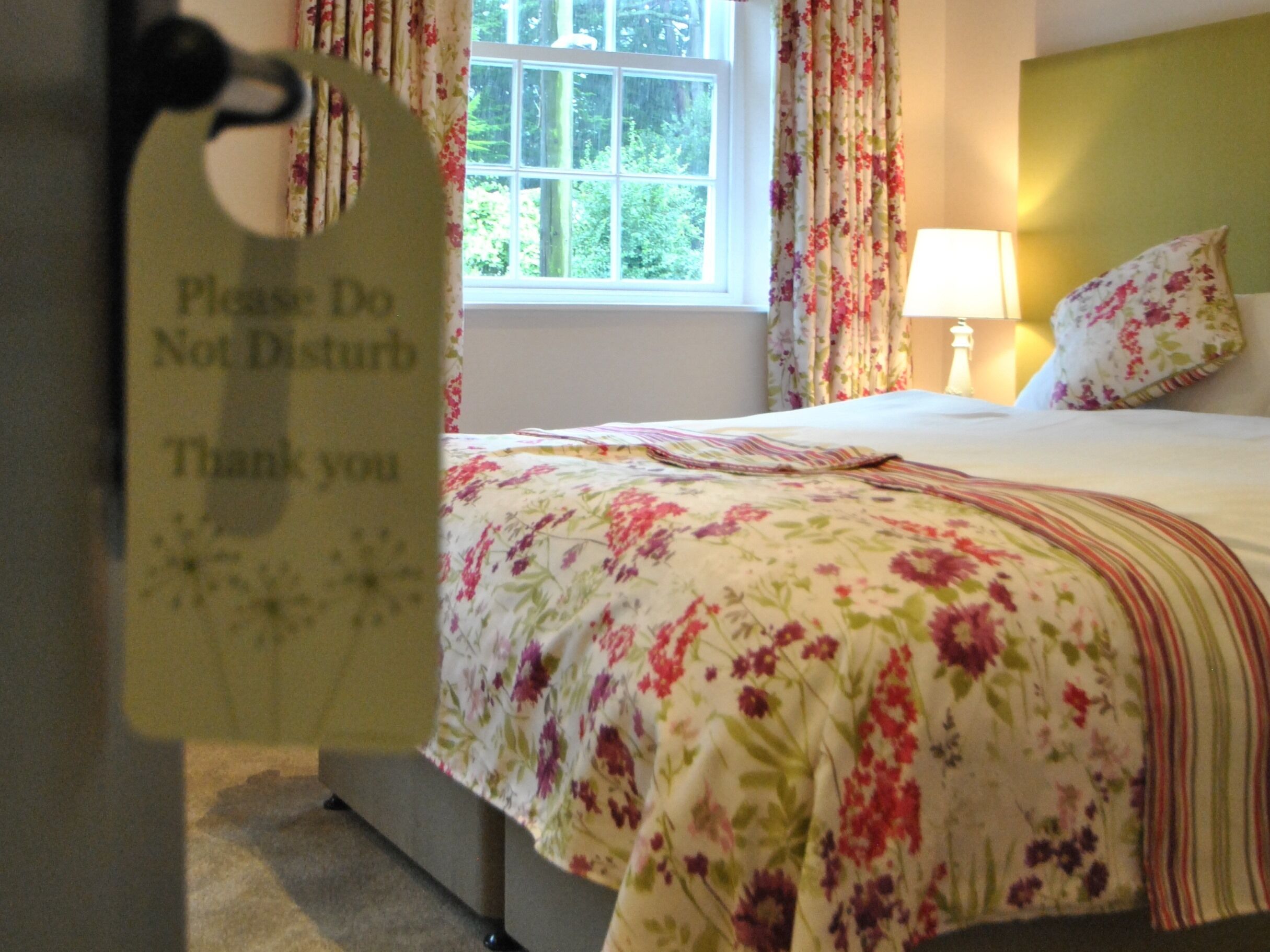 A bedroom with a floral bedspread, a bedside lamp, and a window with curtains.