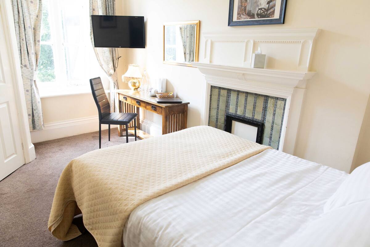 Een hotelkamer met een bed, een bureau met stoel, een televisie, een open haard en een raam met gordijnen.