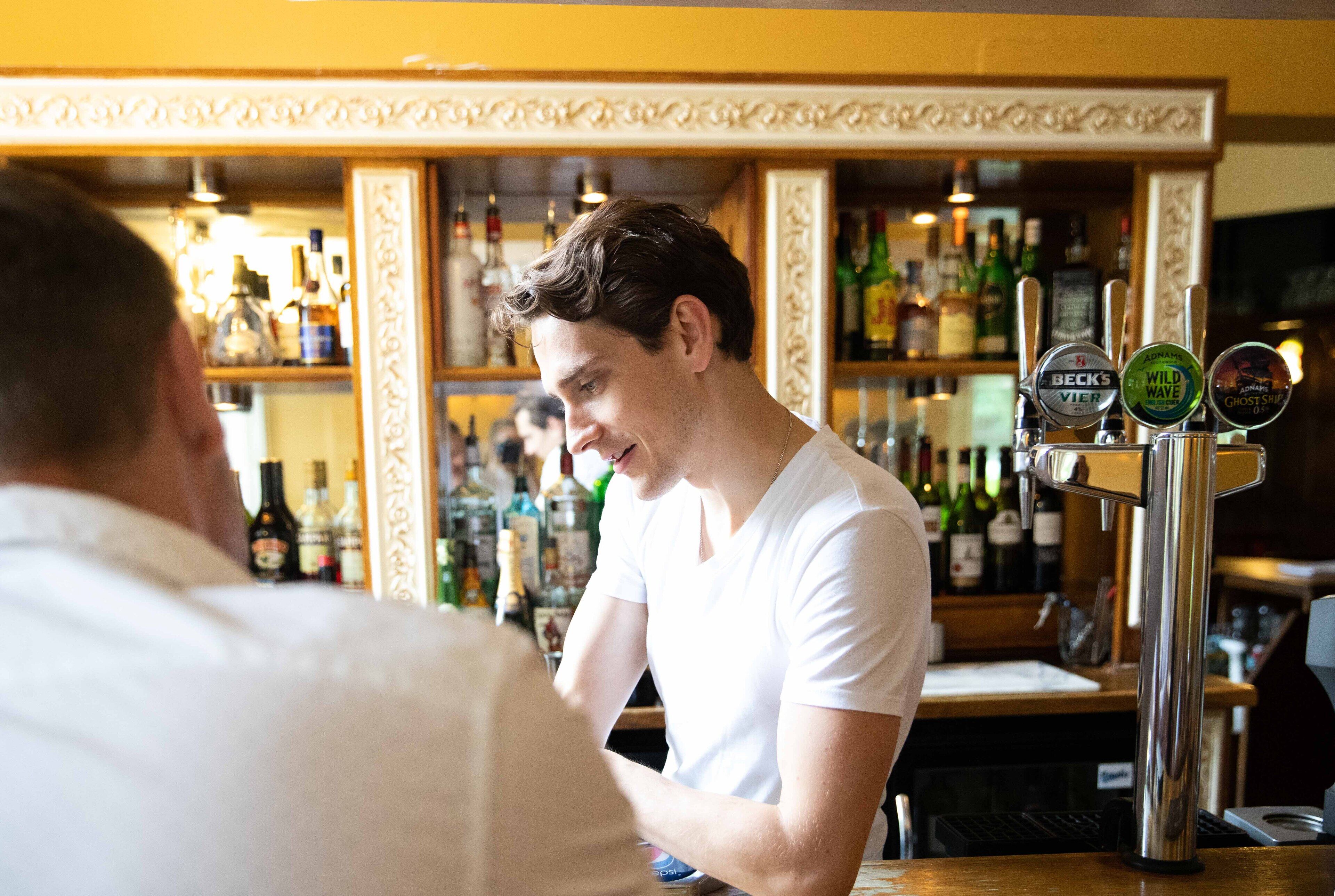 Zona de bar con un bartender atendiendo a un cliente. Hay grifos de cerveza con las etiquetas 'BECK'S VIER', 'WILD WAVE' y 'GHOST SHOT'. El bar tiene estantes con varias botellas y un espejo sobre la barra.