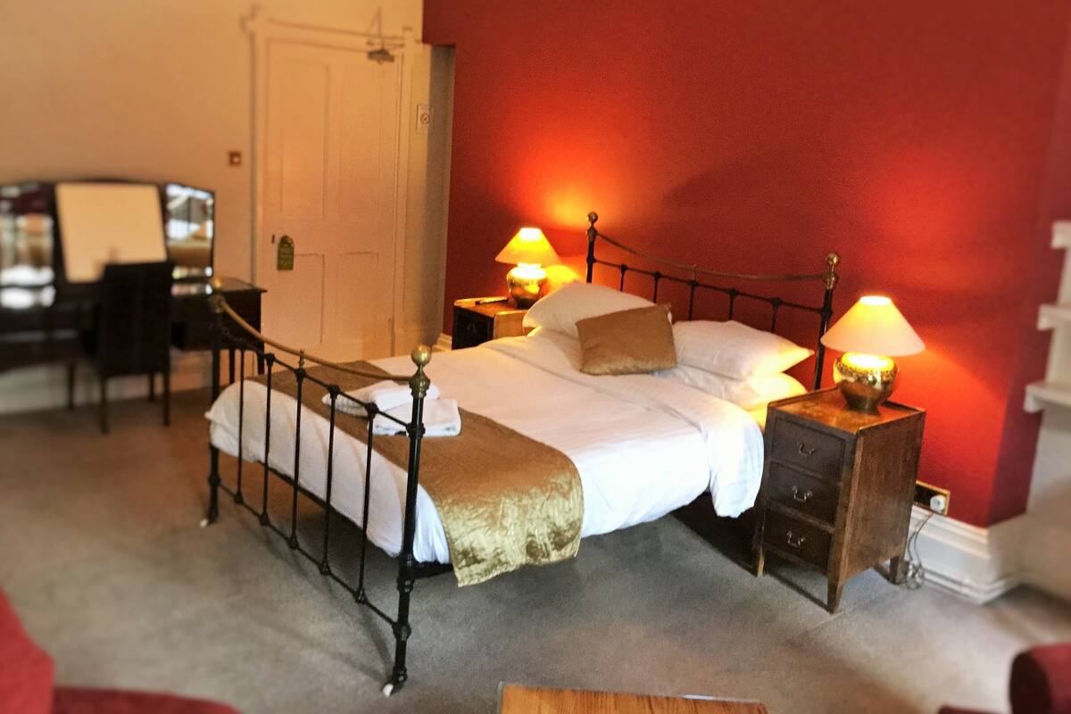 Een netjes opgemaakt bed met witte lakens en een bruin plaid, twee bedlampen, een houten nachtkastje en een bureau met stoel in een kamer met rode wanden.