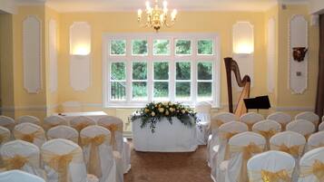 A room with rows of chairs with white covers and gold sashes, a table with a floral arrangement, a harp, and a window with a view of greenery.