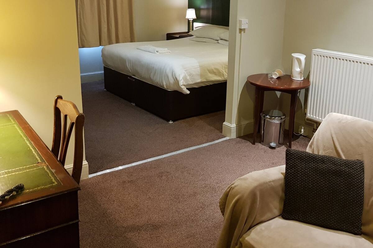 Een hotelkamer met een bed, een tafeltje, een stoel, een radiator en een raam met gordijnen.