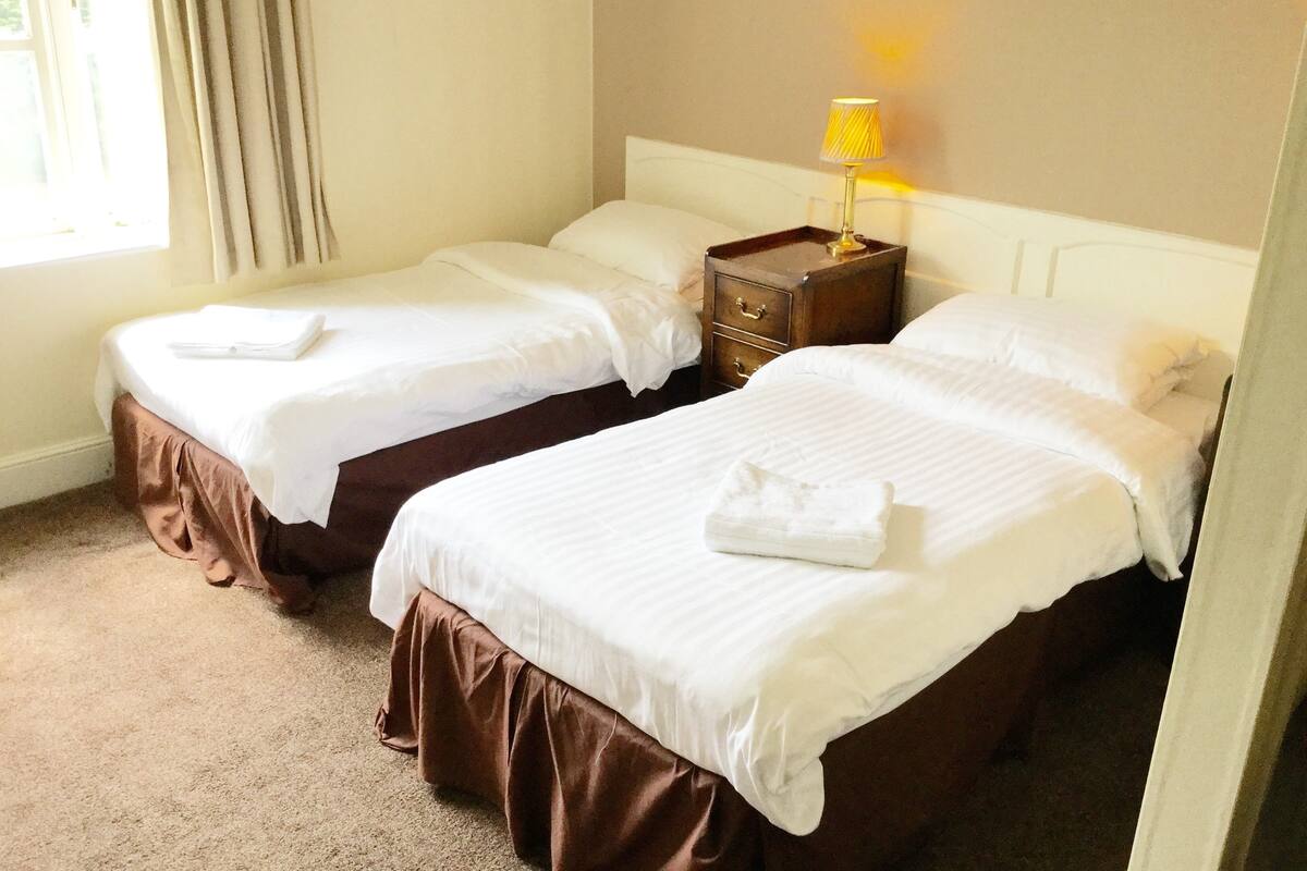 Een hotelkamer met twee bedden, een nachtkastje met een lamp en een raam met gordijnen.