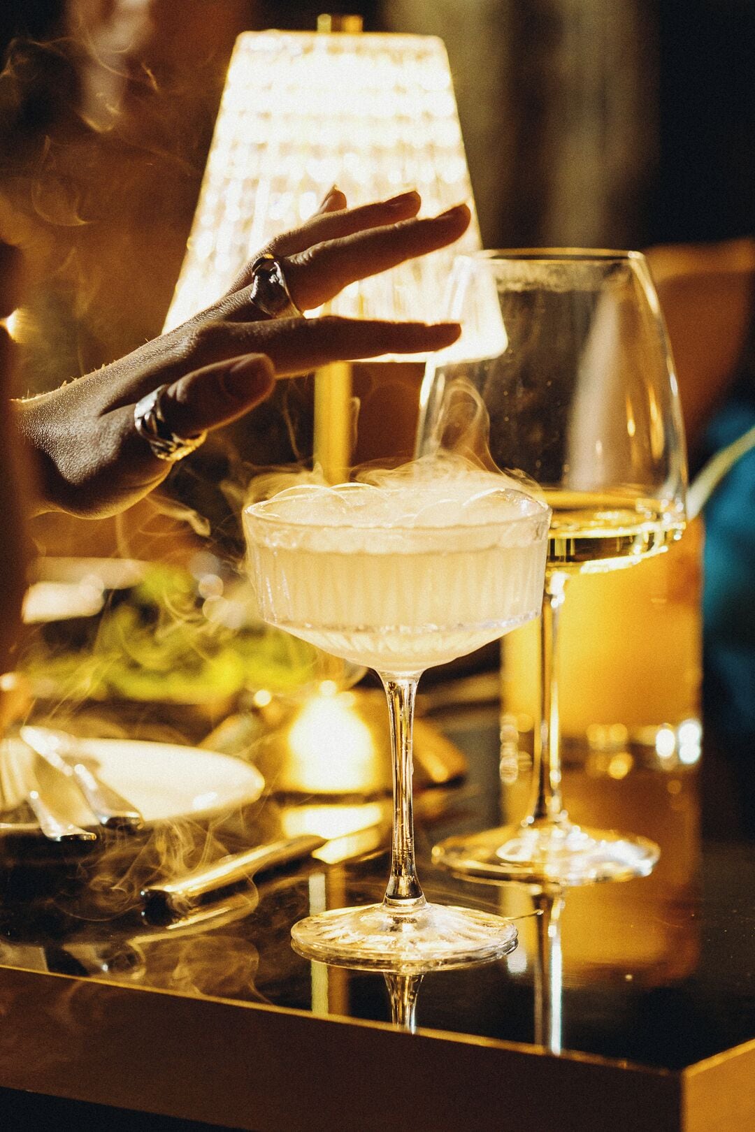 Un bar con una copa de vino blanco y un cóctel con hielo, una mano alcanzando la bebida, y una lámpara al fondo.