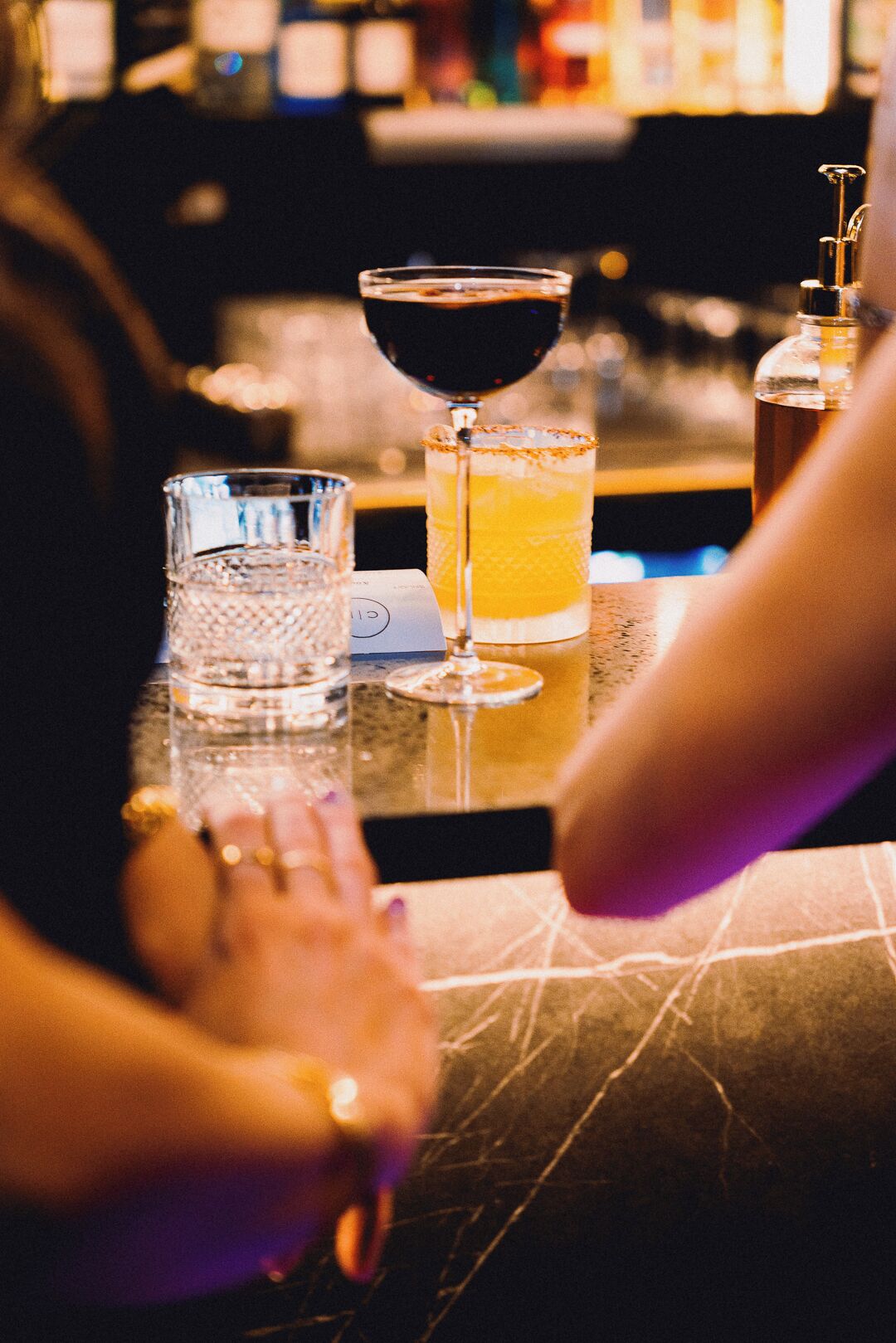 Eine Theke mit einem Glas dunkler Flüssigkeit, einem Glas mit einem gelblichen Getränk und einem klaren Wasserglas.