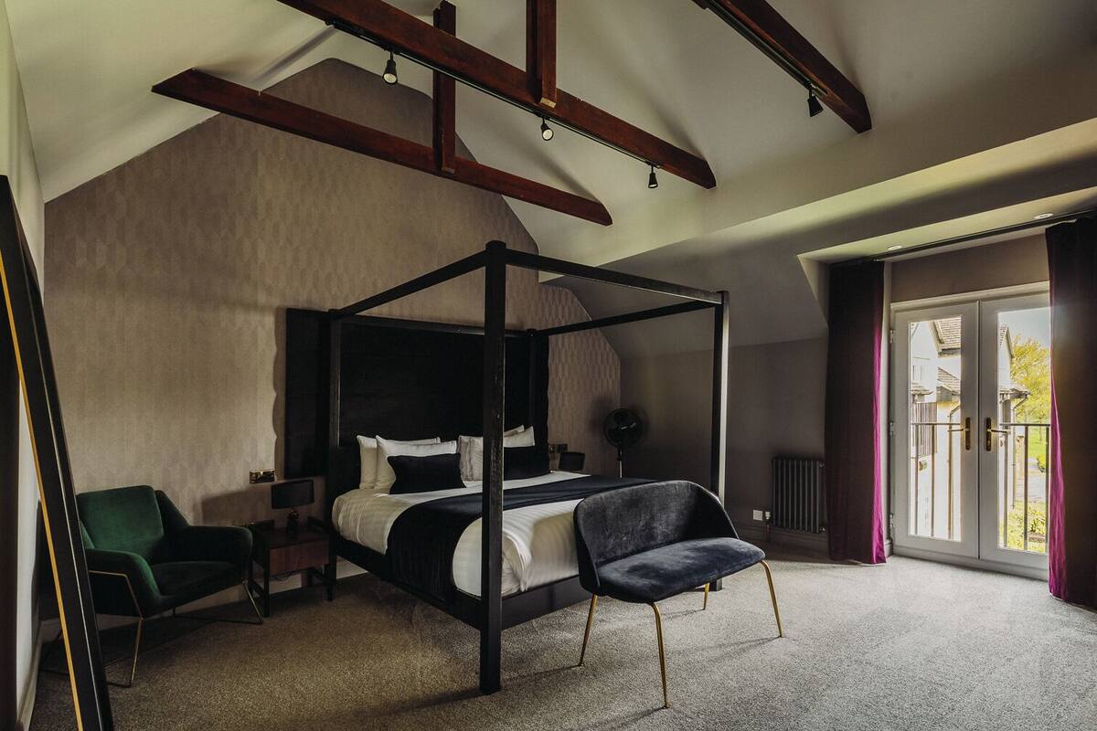 A four-poster bed with a dark headboard, a green armchair, a black chair, and a bedside table with a lamp.