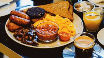 A breakfast table with plates of food, including toast, sausages, beans, scrambled eggs, and a glass of orange juice.