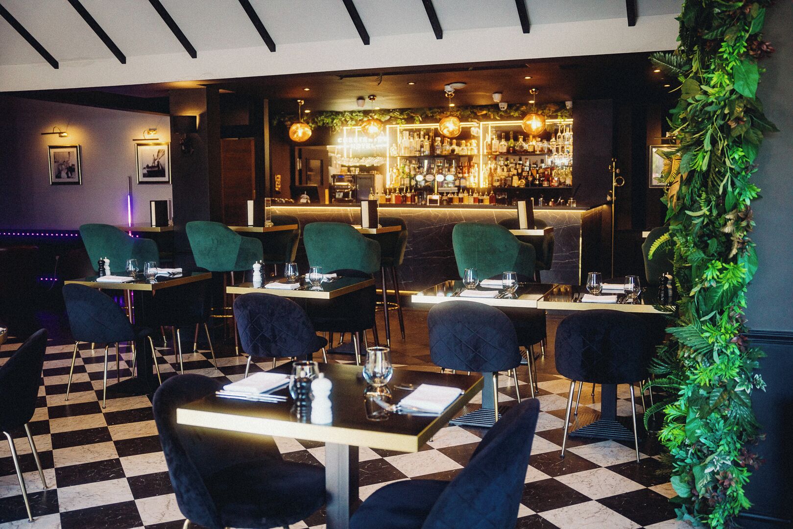 A restaurant with checkered flooring, green upholstered chairs, and a bar area with a variety of bottles and glasses.