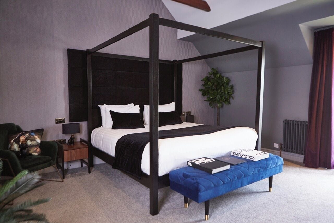 A four-poster bed with a dark frame, a bedside table with a lamp, a blue bench, and a book titled 'TOM FORD 002' on the bench.