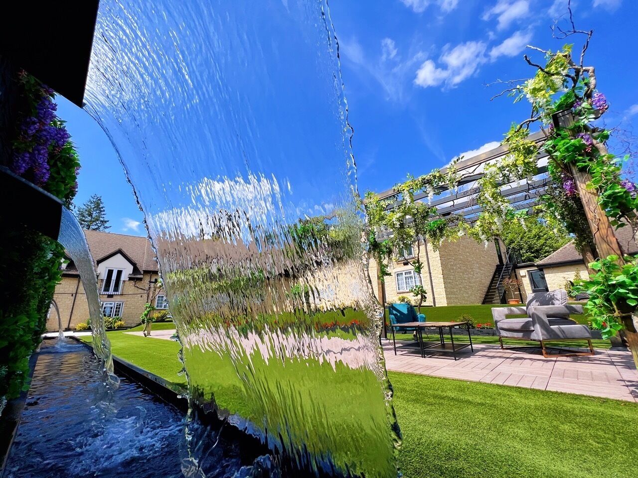 A garden with a water feature, a lawn, a patio with outdoor furniture, and a building in the background.