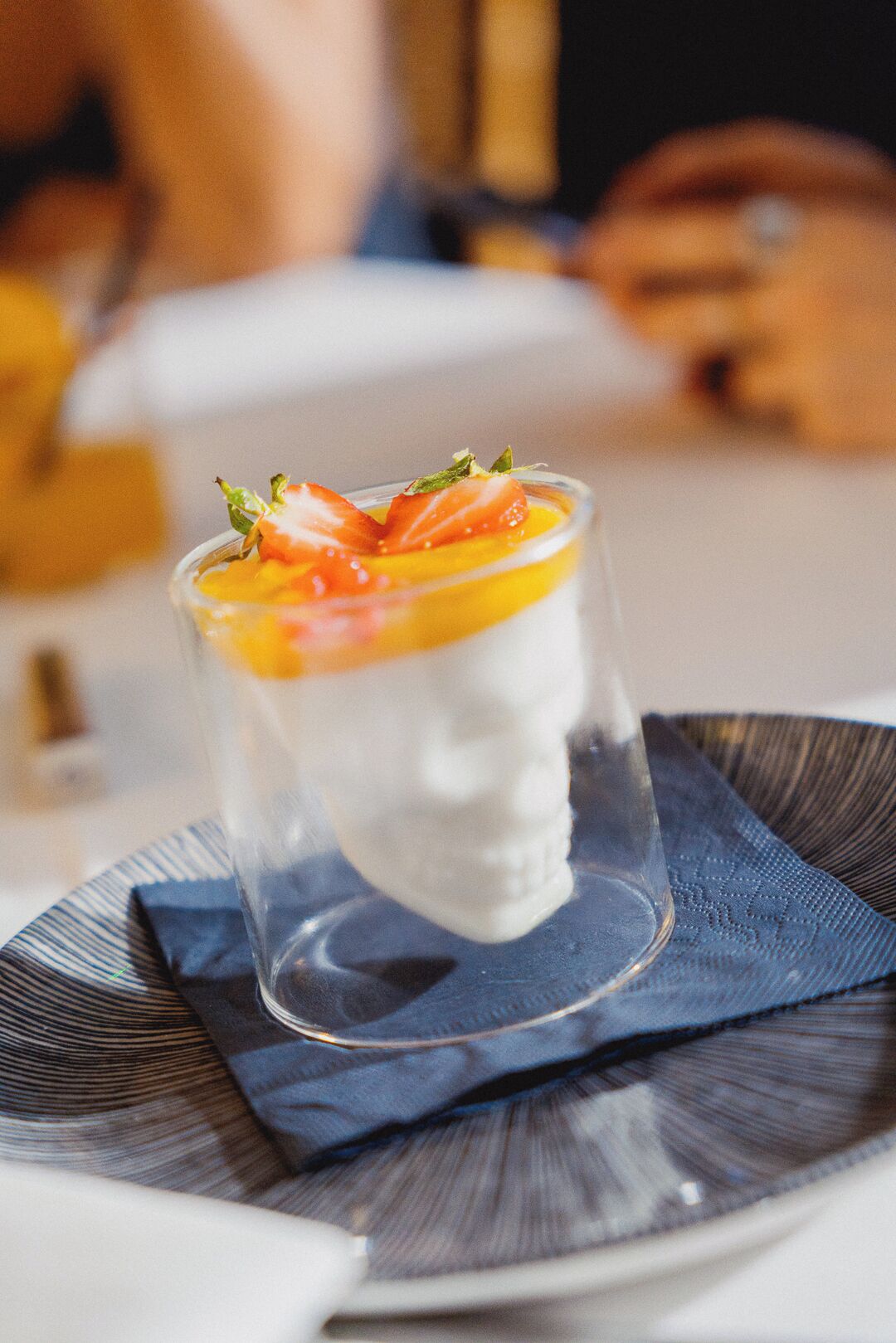 Un postre en un vaso transparente sobre un plato con un mantelito azul debajo.