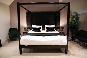 Una cama con dosel con ropa de cama blanca y almohadas negras, flanqueada por dos mesitas de noche con lámparas, en una habitación con una silla verde y un sofá negro.