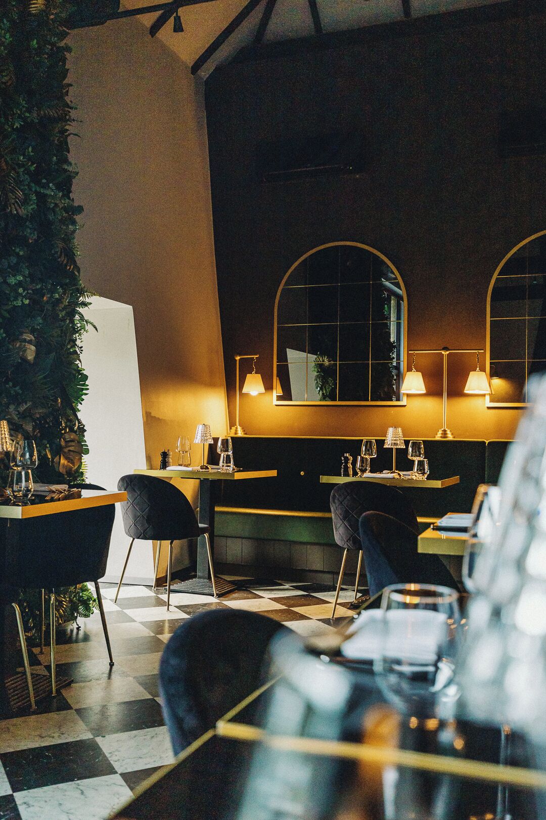 Interior de un restaurante con suelo a cuadros, sillas de tonos oscuros y mesas dispuestas con copas de vino y cubiertos.