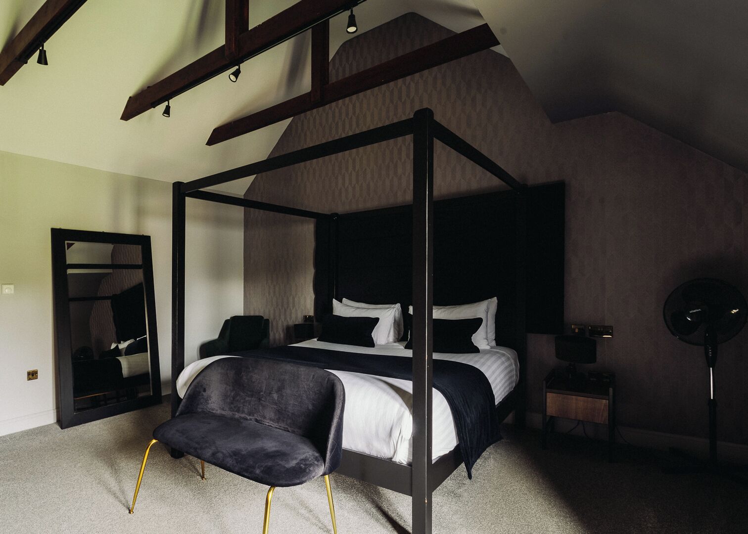 A four-poster bed with a dark frame and white bedding, a black upholstered chair with gold legs, a bedside table with a lamp, and a large mirror on the wall.