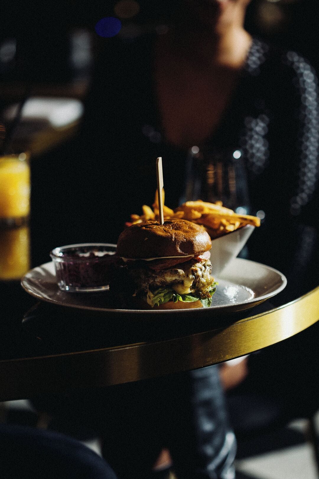 Un hamburger, maintenu par un cure-dent, est servi sur une assiette, accompagné de frites et d’un petit bol de sauce, sur une table dans un endroit à la lumière tamisée.