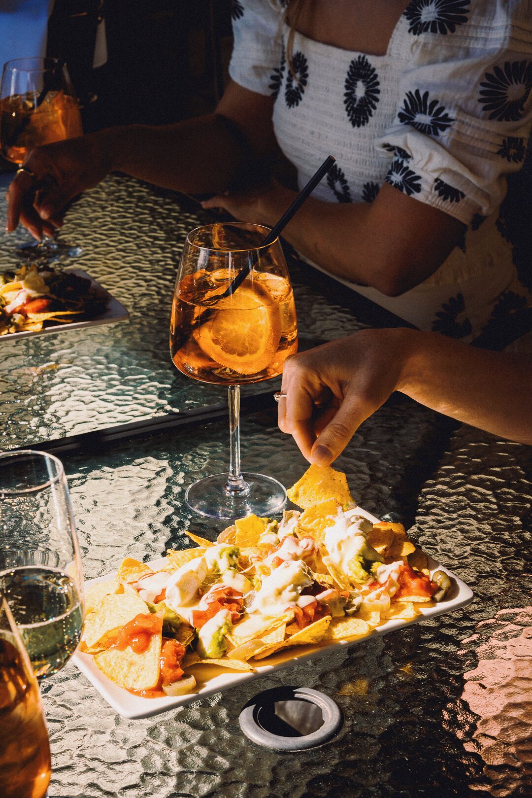 A dining table with a plate of nachos, a glass of iced beverage, and a wine glass.