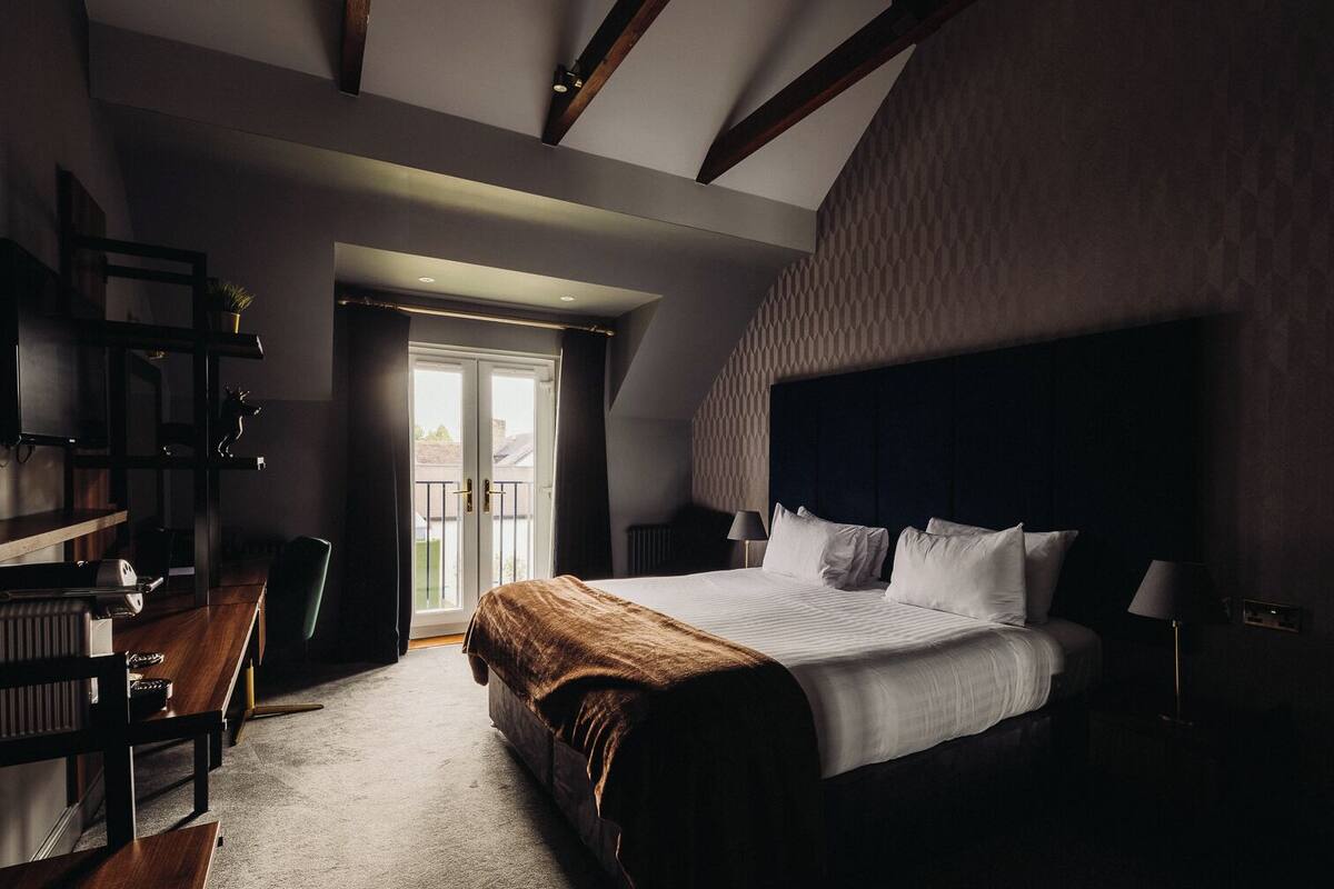 A hotel room with a large bed, a desk, a chair, and a view of the outdoors through a sliding door.