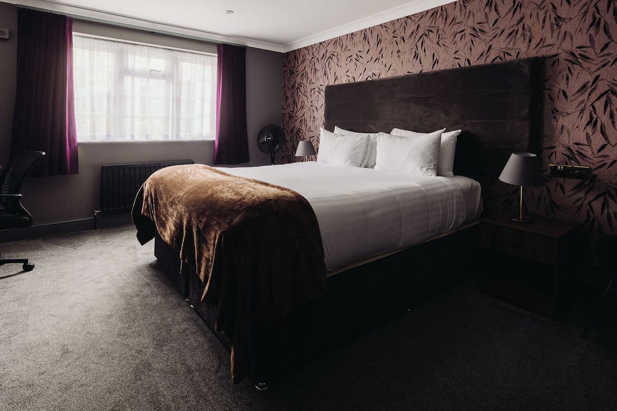 A hotel room with a large bed, a bedside table with a lamp, a chair, and a radiator.