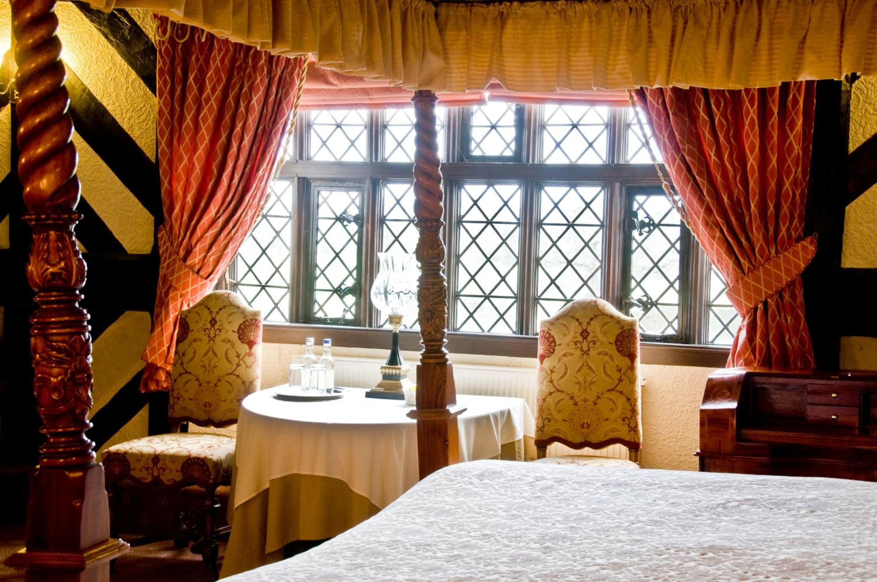 A four-poster bed with a canopy, two upholstered chairs, a round table with a white tablecloth, and a window with a lattice design.