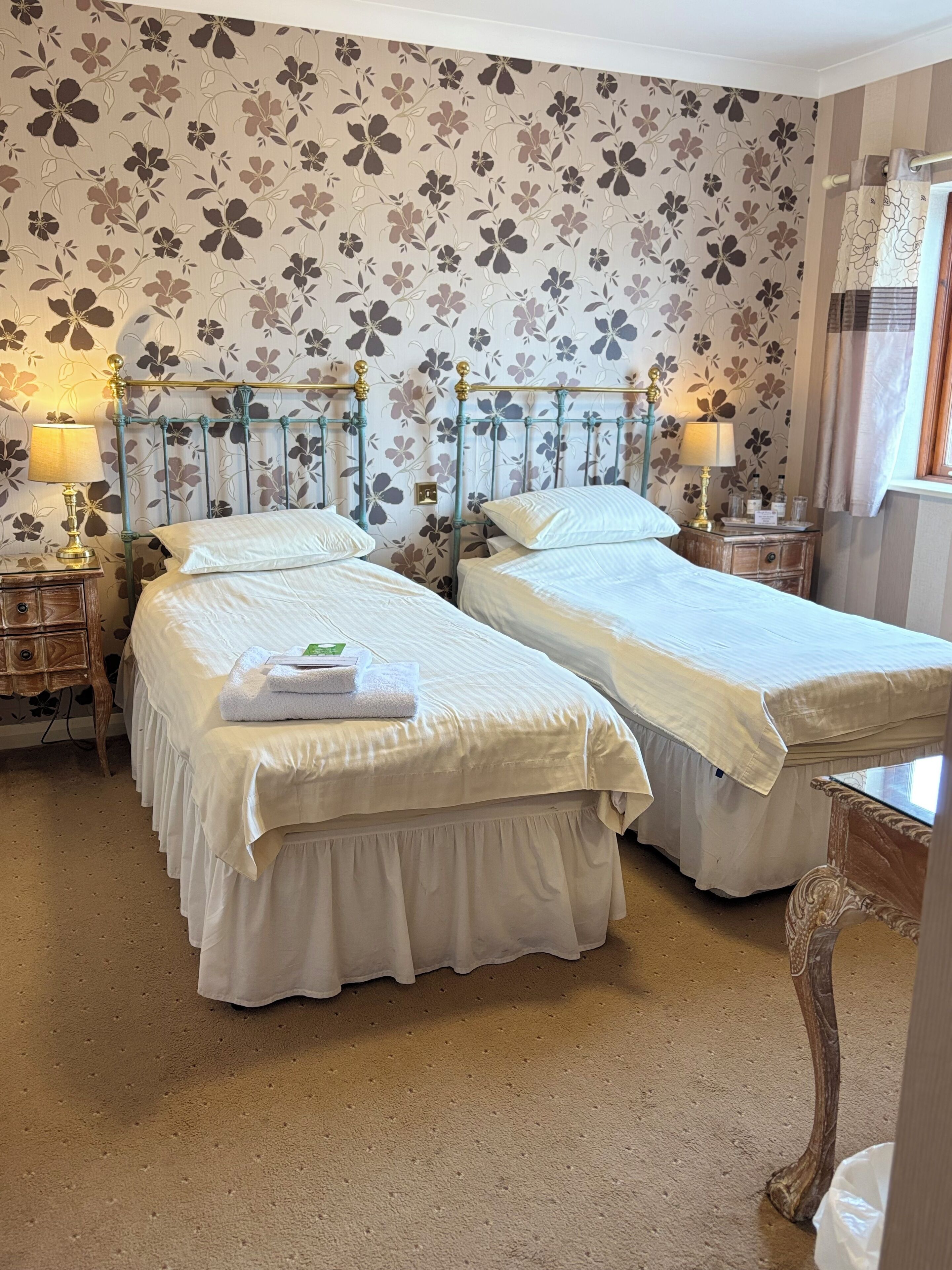 A hotel room with two beds, floral wallpaper, and a classic metal bed frame.