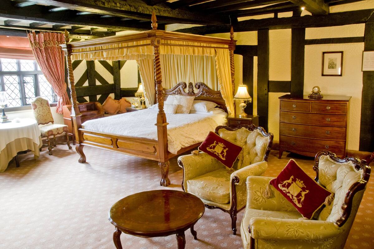A four-poster canopy bed with matching armchairs, a wooden nightstand, and a small table in a traditional room with exposed beams and a patterned carpet.
