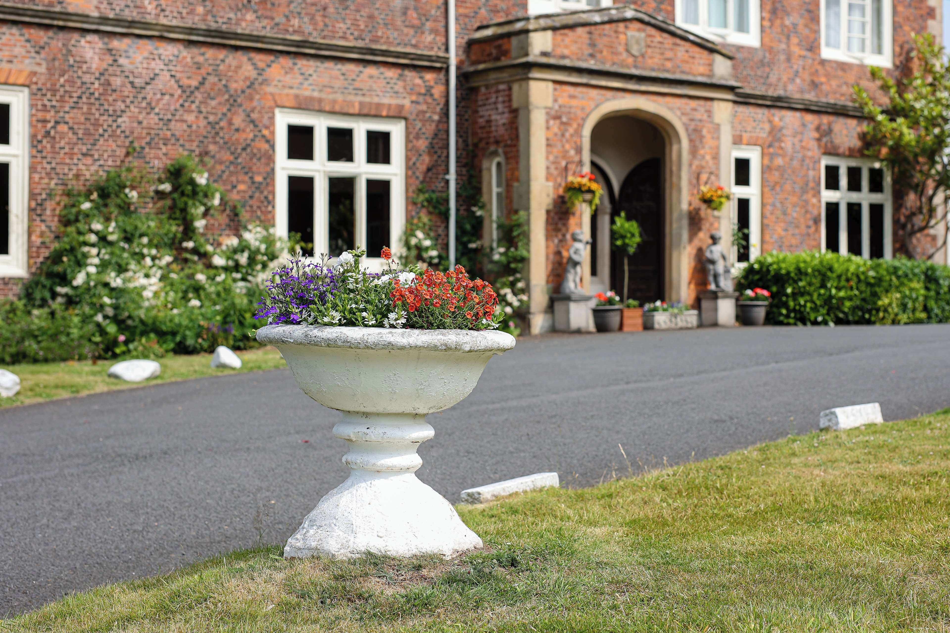 A white urn with flowers in front of a brick building with a large arched entrance.
