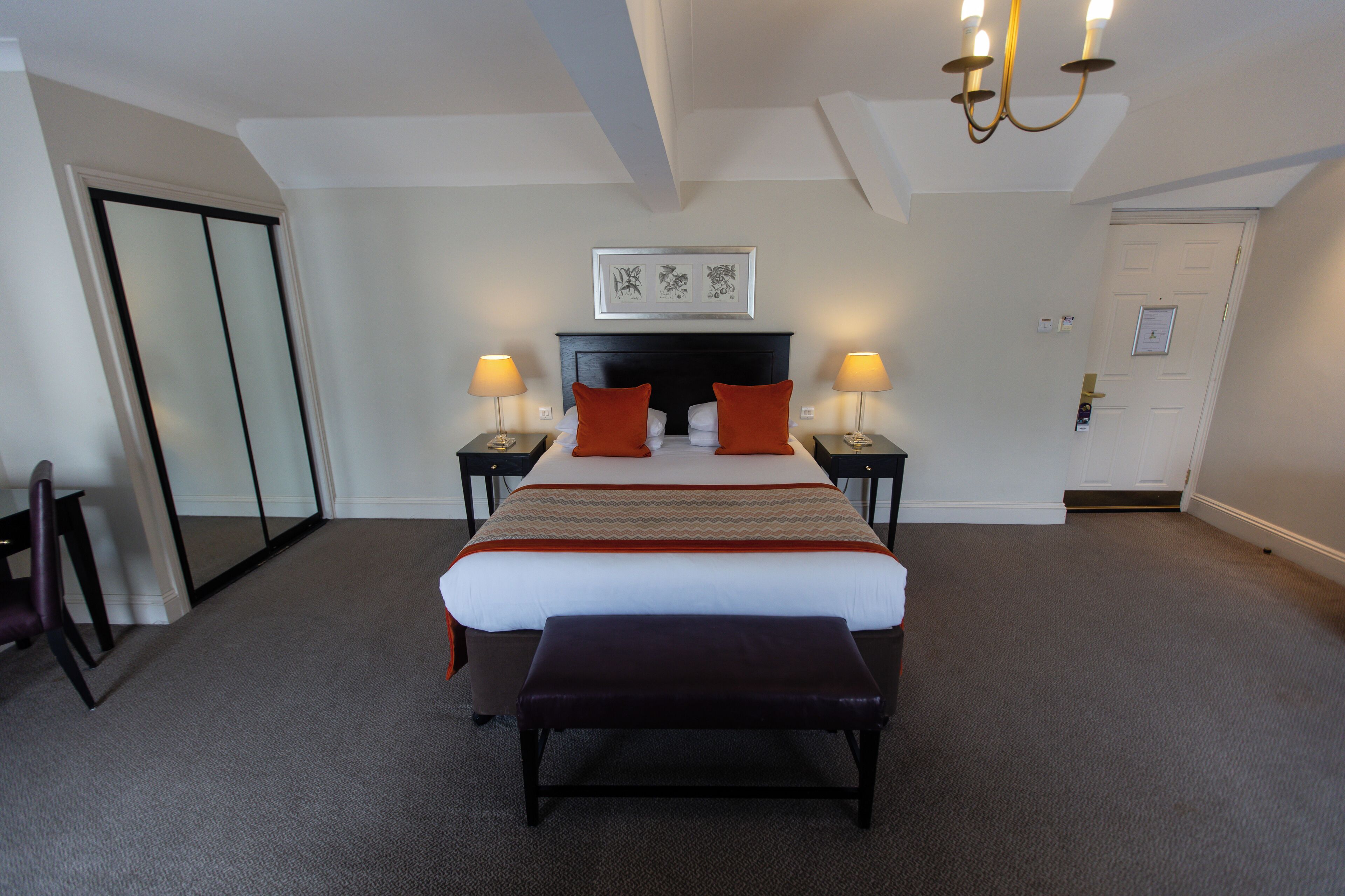 A hotel room with a large bed, two bedside tables with lamps, a bench at the foot of the bed, a door, and a framed picture on the wall.