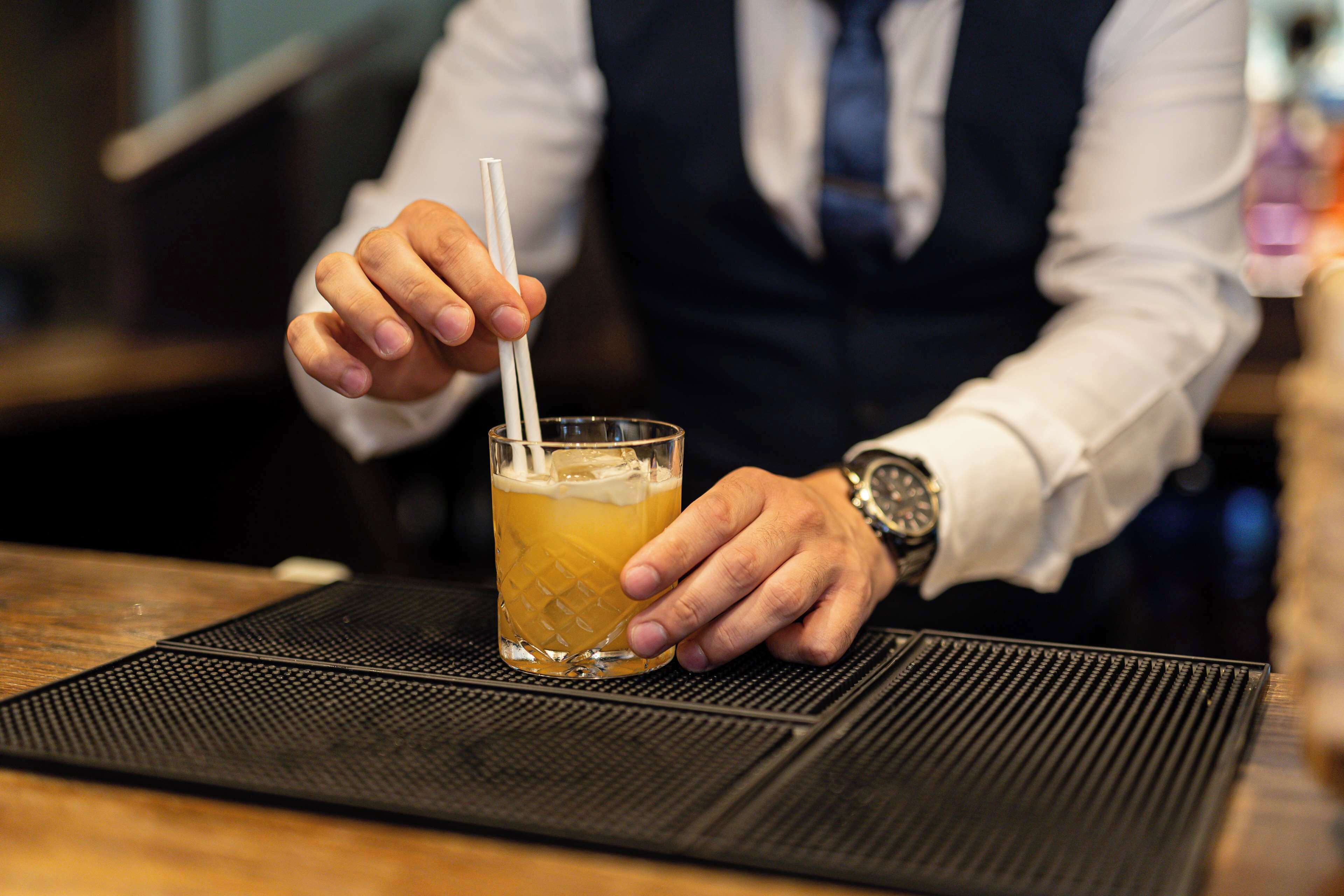 Eine Person hält ein Glas mit Trinkhalm, trägt eine Armbanduhr und steht an einer Bar.