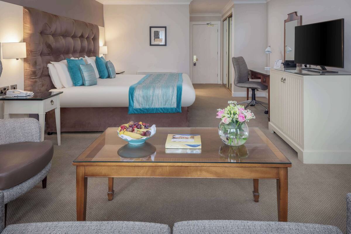 Une chambre d’hôtel avec un grand lit, un bureau avec un ordinateur, une télévision et une table basse sur laquelle se trouvent un panier de fruits et un vase de fleurs.