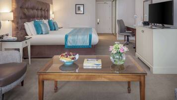 Une chambre d’hôtel avec un grand lit, un bureau avec un ordinateur, une télévision et une table basse sur laquelle se trouvent un panier de fruits et un vase de fleurs.
