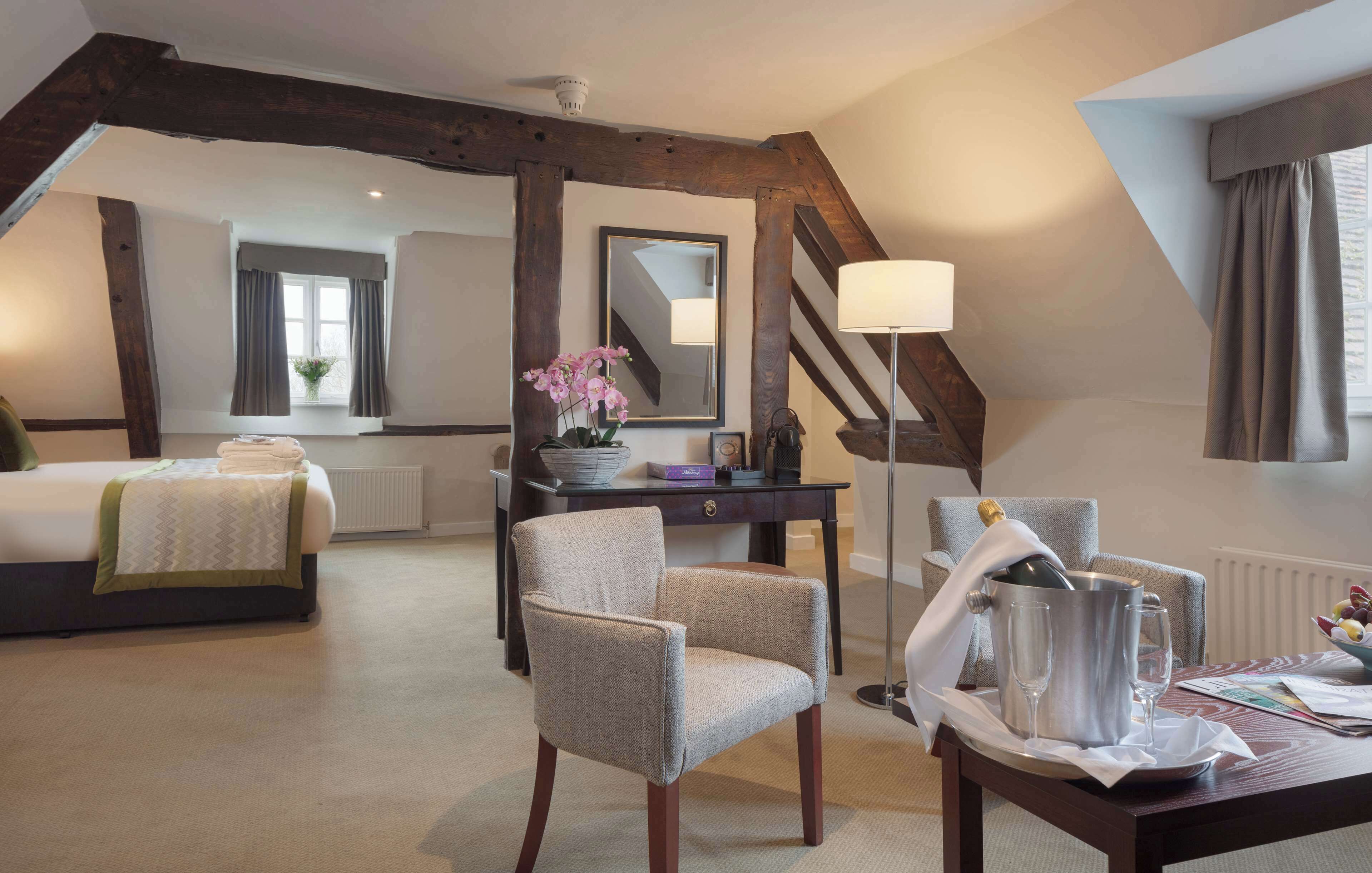 Une chambre d’hôtel avec un lit, un bureau avec une lampe, une chaise et une table sur laquelle se trouvent une bouteille et des verres.