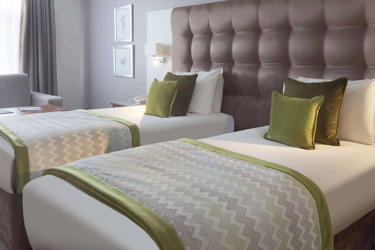 Une chambre d’hôtel avec deux lits, chacun recouvert d’une couvre-lit à motifs en zigzag vert et blanc, doté d’une tête de lit capitonnée et agrémenté d’une table de chevet avec une lampe.