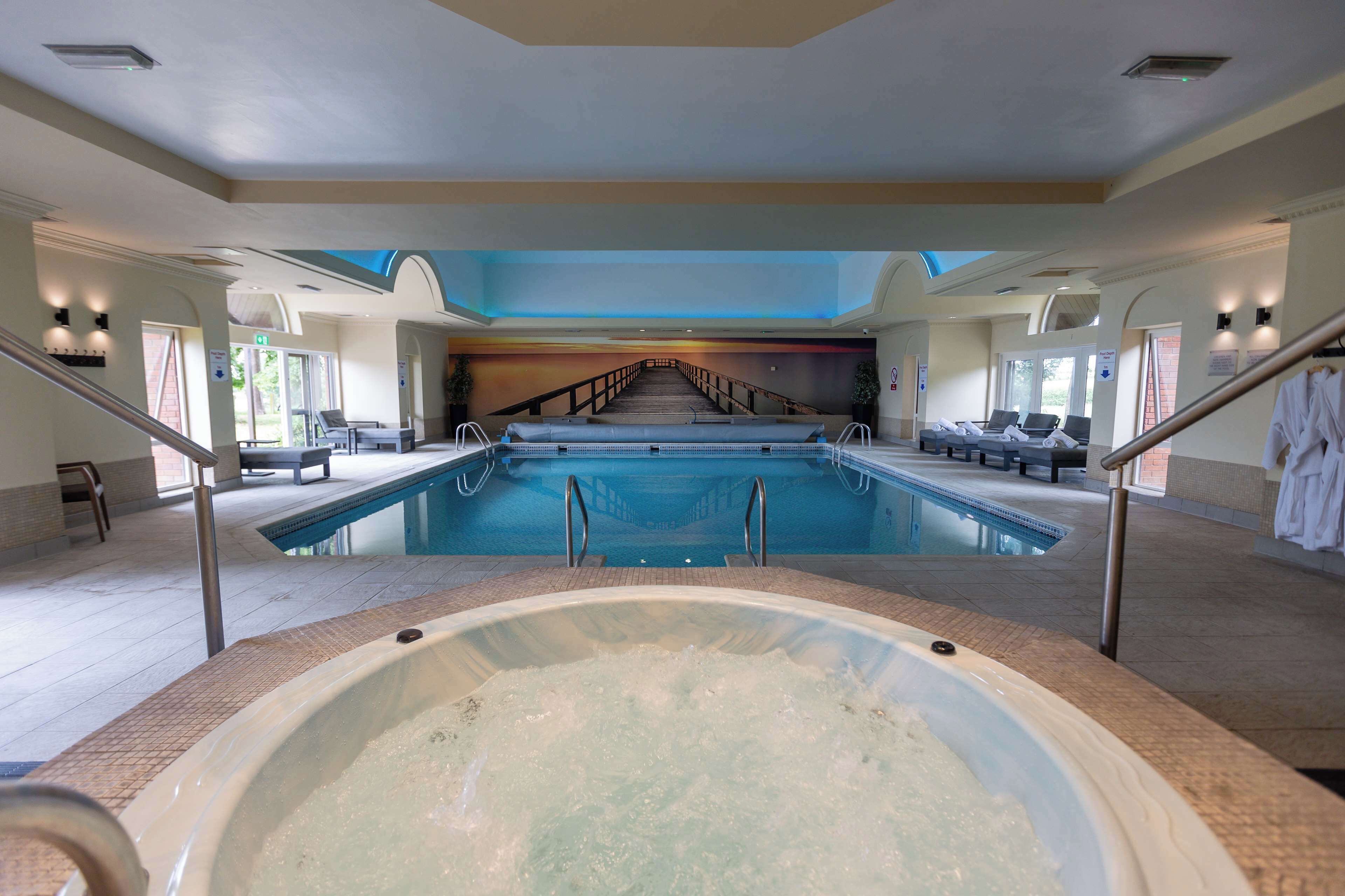 A large indoor swimming pool with a hot tub in the foreground, a staircase leading to a second level, and lounge chairs along the poolside.