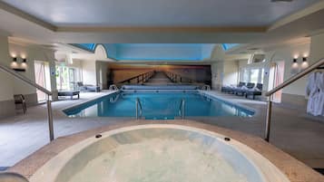 A large indoor swimming pool with a hot tub in the foreground, a staircase leading to a second level, and lounge chairs along the poolside.