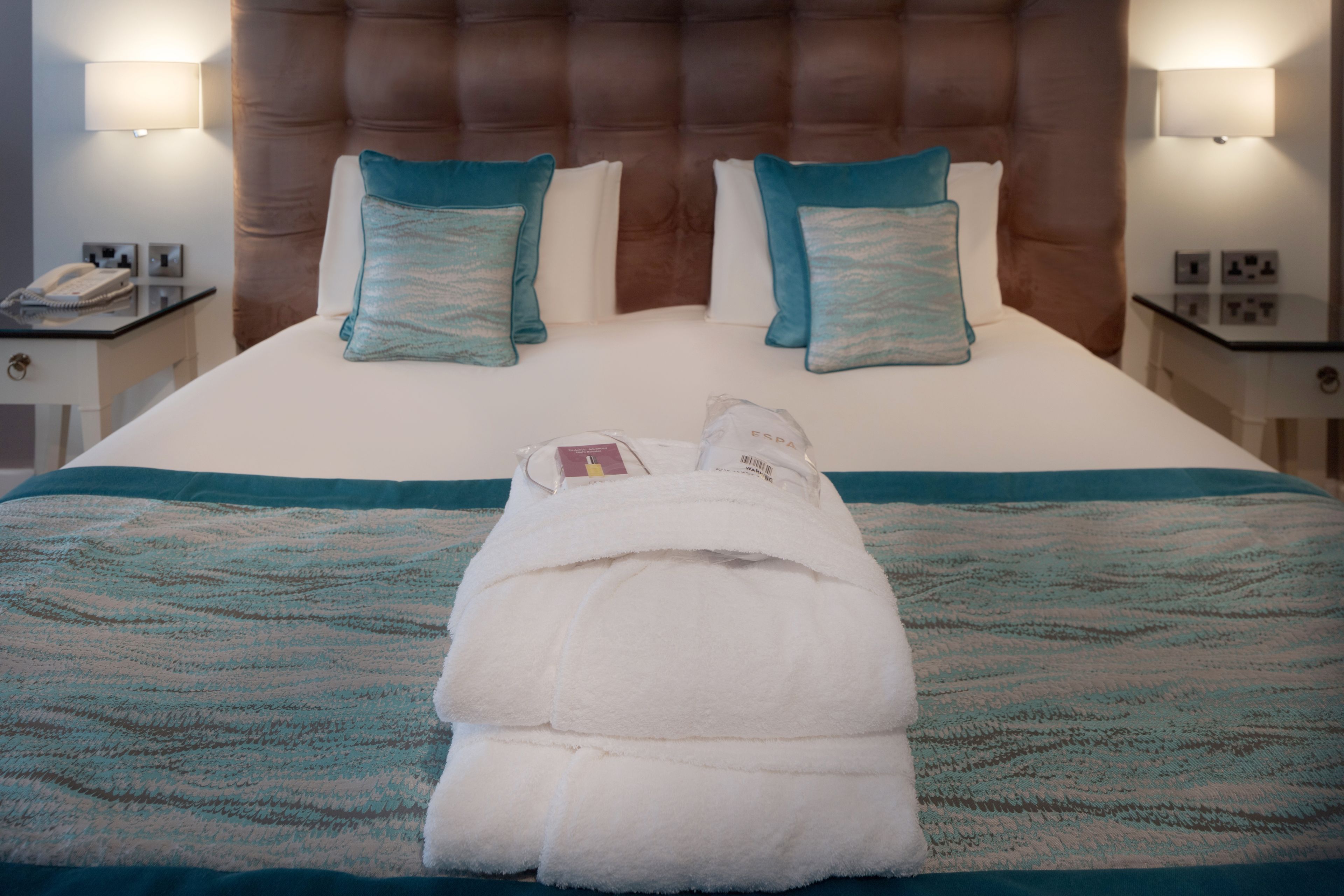 A neatly made bed with a folded towel, two pillows, and a bedside table with a lamp and a phone.