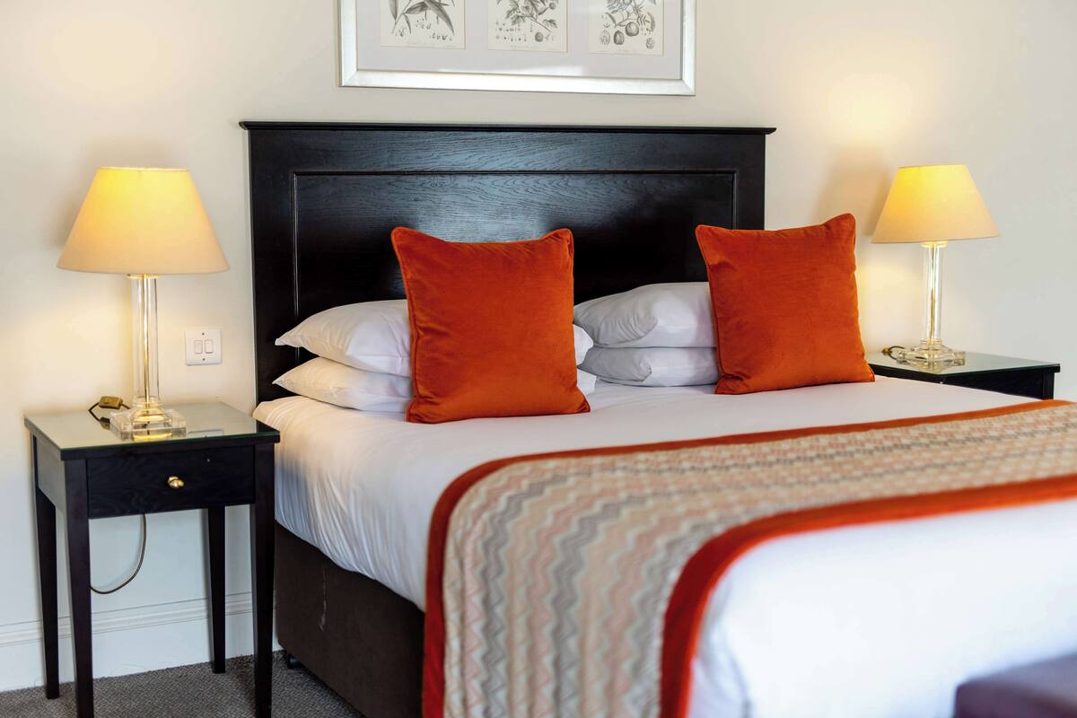 Un lit bien fait, avec du linge de lit blanc et deux oreillers orange, flanqué de deux tables de chevet avec des lampes, le tout placé devant une tête de lit en bois foncé.