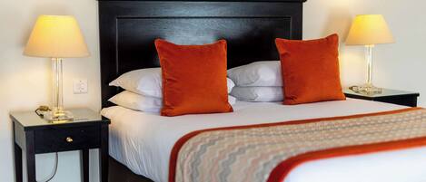 A neatly made bed with white linens and two orange pillows, flanked by two bedside tables with lamps, set against a dark wooden headboard.
