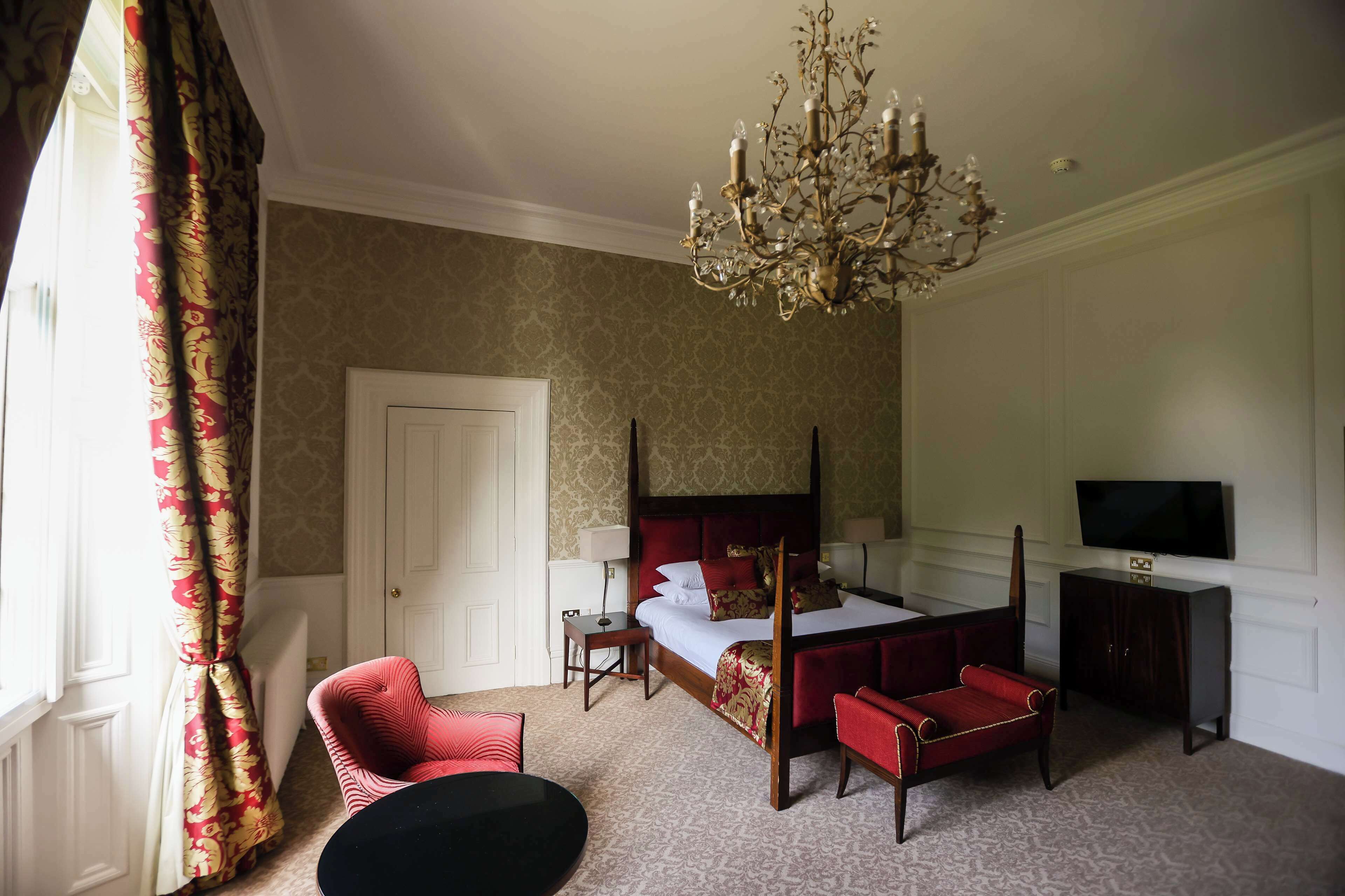 A four-poster bed with a television on a stand, a red upholstered chair, and a chandelier.