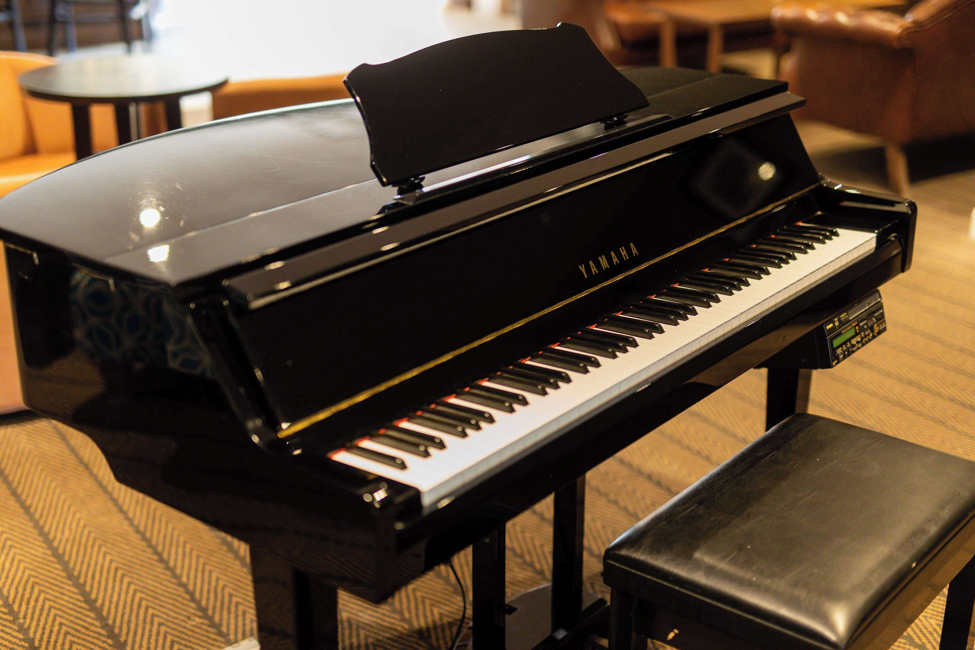 Un piano de cola Yamaha negro con un banco, ubicado en una habitación con alfombra estampada y una silla de cuero marrón al fondo.