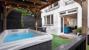 Relax in a shaded hot tub area surrounded by a well-maintained lawn and patio with outdoor furniture.