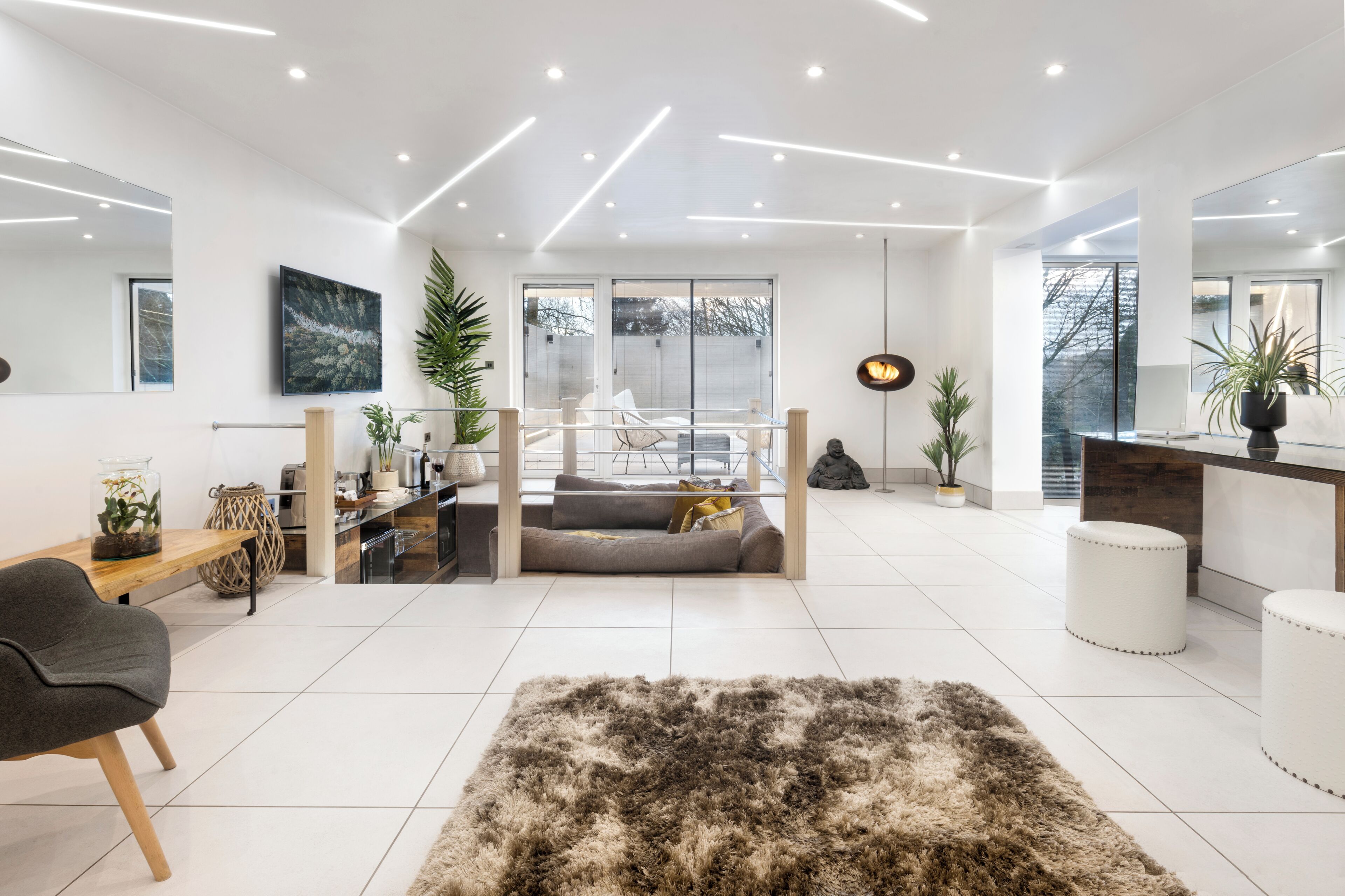 A modern living room with a sofa, a coffee table, and a rug. There are plants, a dining table, and a kitchen area visible.