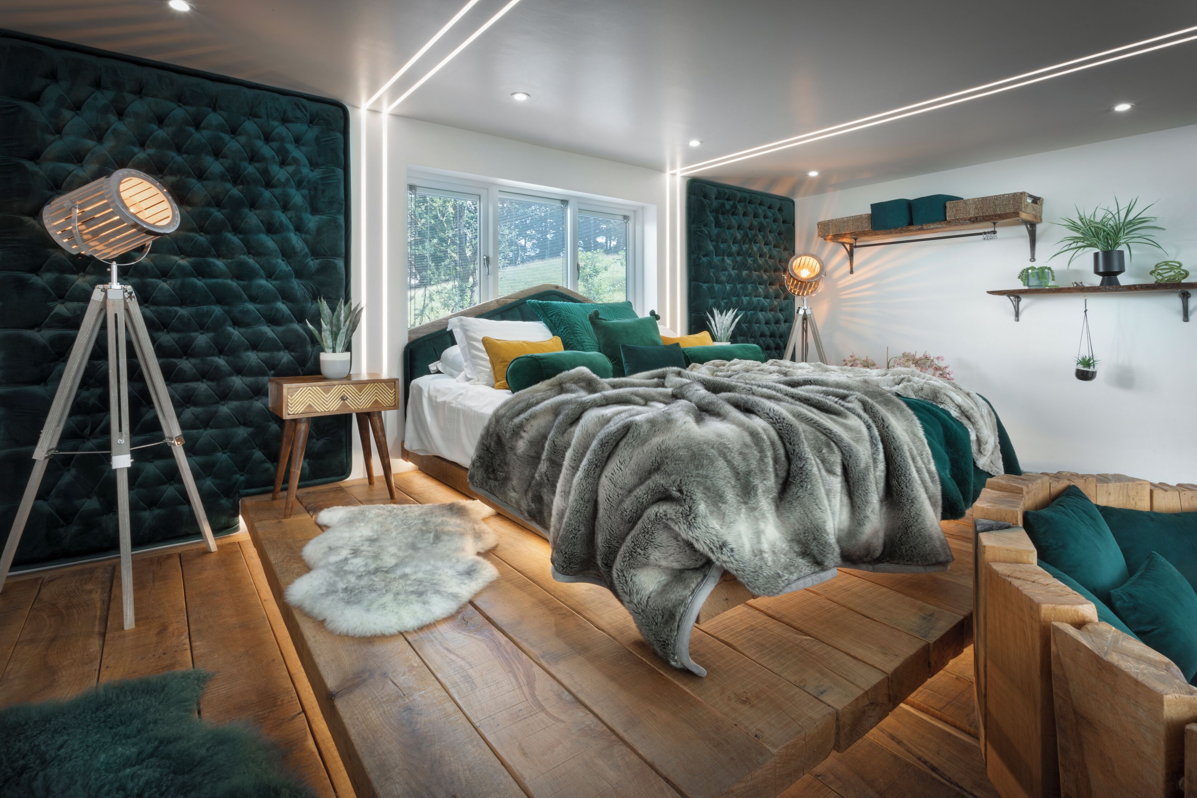 A modern bedroom with a tufted headboard, a wooden bed frame, a large grey fur throw, a floor lamp, and a view of greenery outside.
