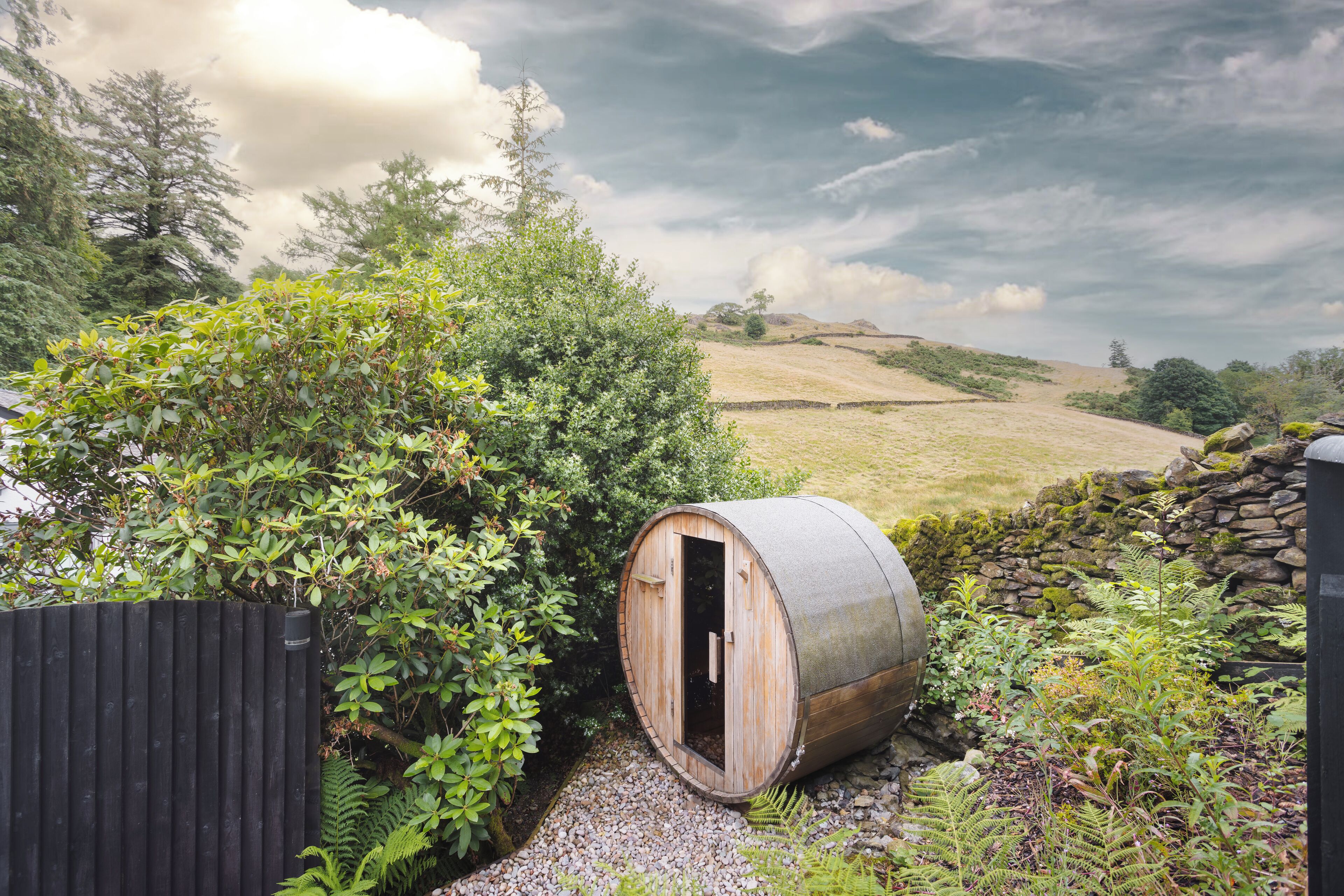 A rustic wooden barrel structure with a door, surrounded by lush greenery and a stone wall, set against a backdrop of rolling hills and a cloudy sky.