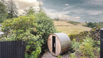 A rustic wooden barrel structure with a door, surrounded by lush greenery and a stone wall, set against a backdrop of rolling hills and a cloudy sky.