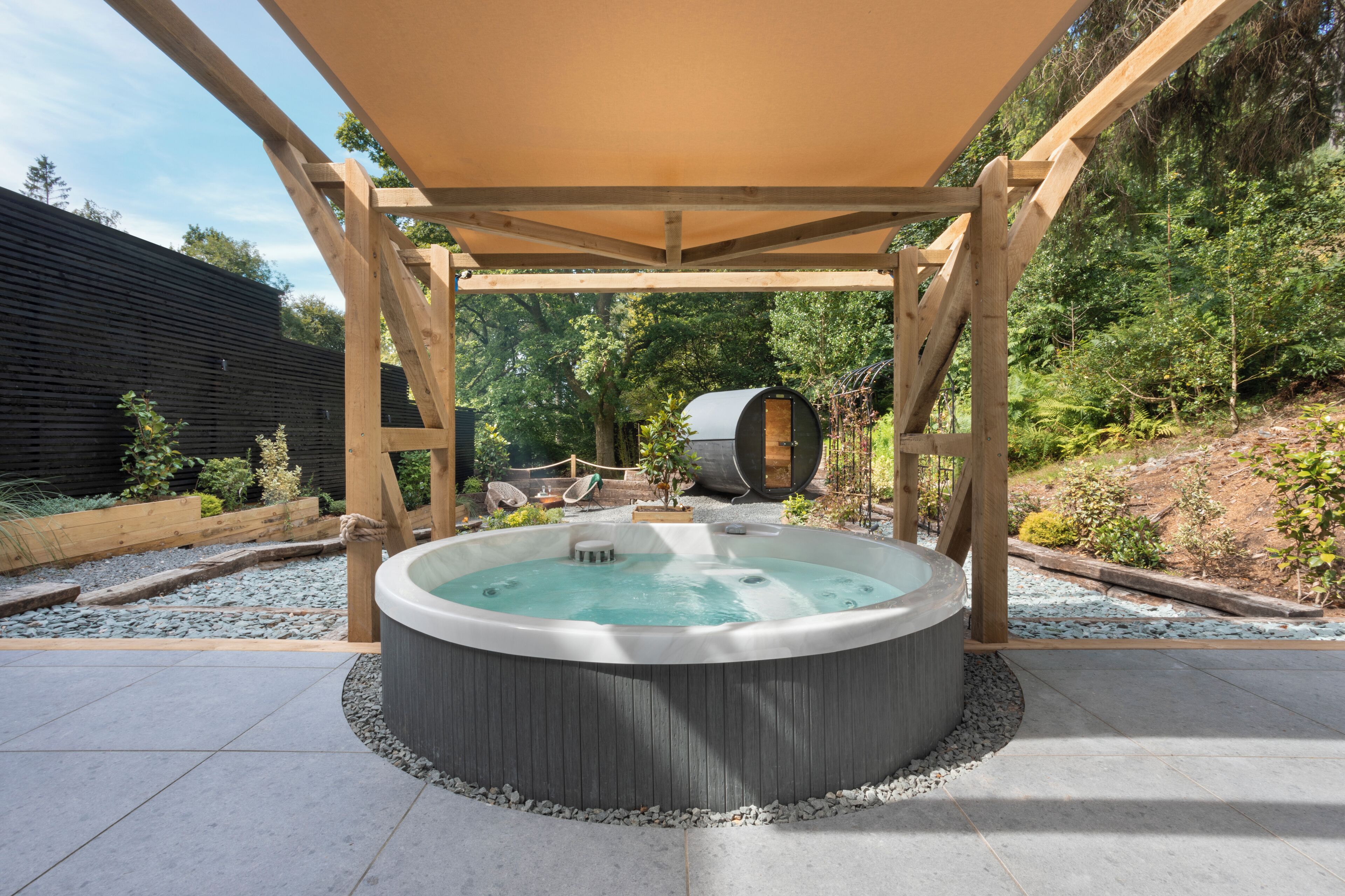 A hot tub under a wooden pergola in a landscaped garden with a wooden fence and trees in the background.