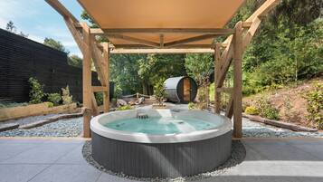 A hot tub under a wooden pergola in a landscaped garden with a wooden fence and trees in the background.