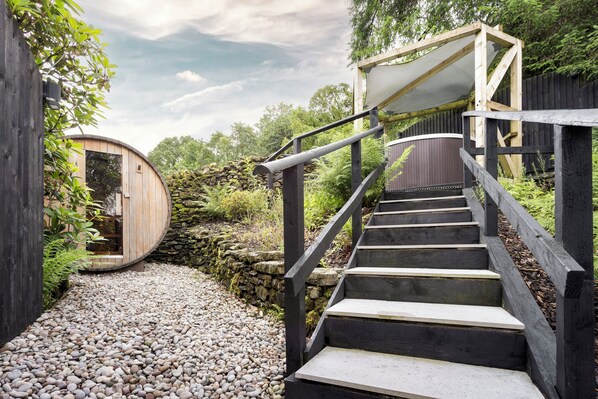 A wooden spiral staircase leading to a covered wooden structure amidst a garden with a stone wall and gravel path. - Aphrodites Group (Windermere)