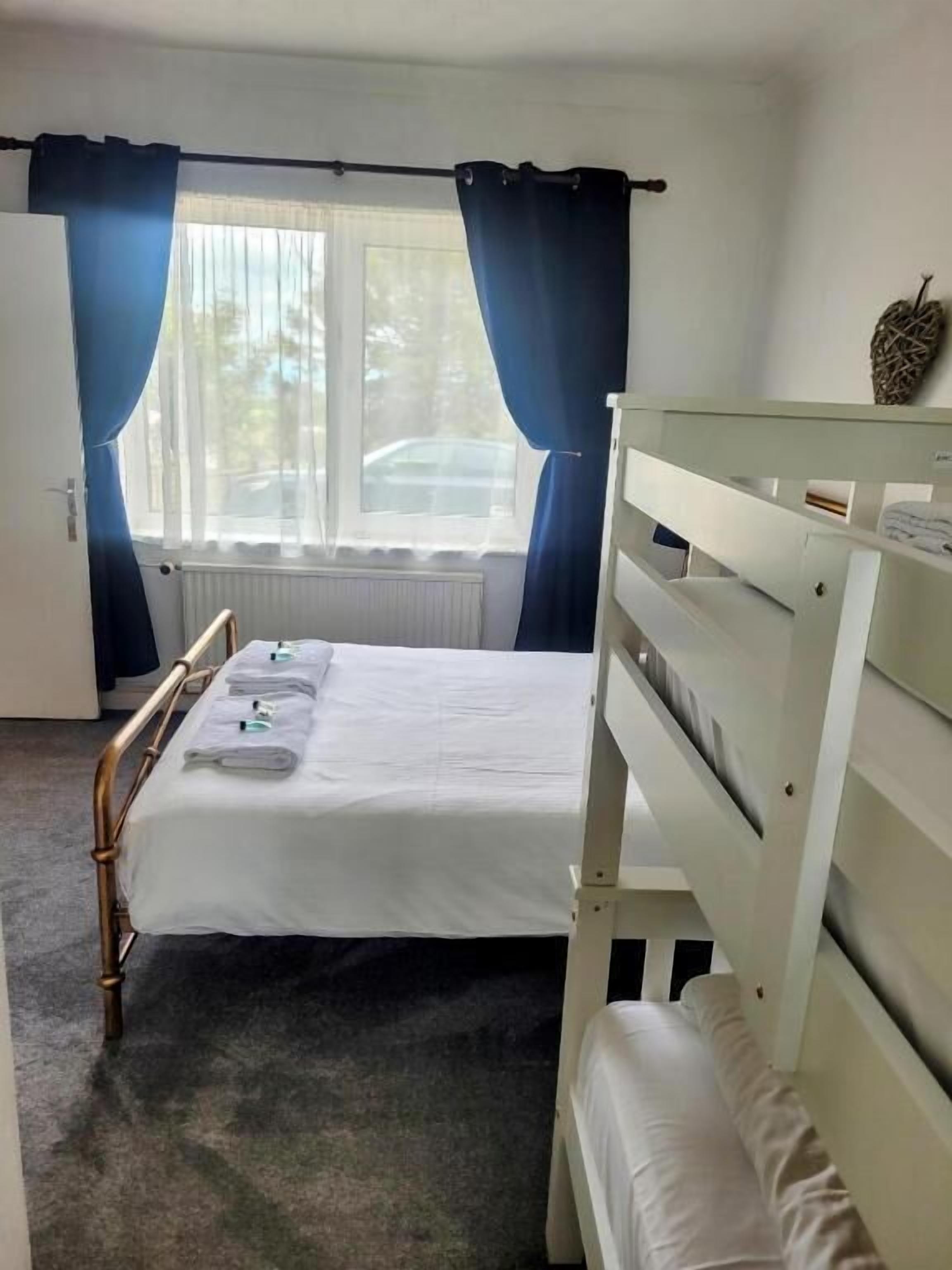A single-bed room with a window, curtains, a radiator, and a small bedside table.