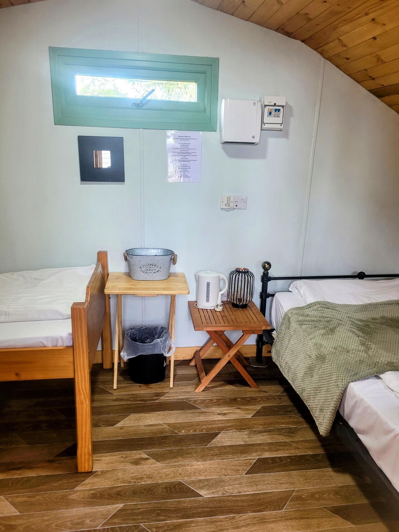 A small, cozy room with two wooden beds, a small table with a kettle, a trash bin, and a window with a view of greenery.